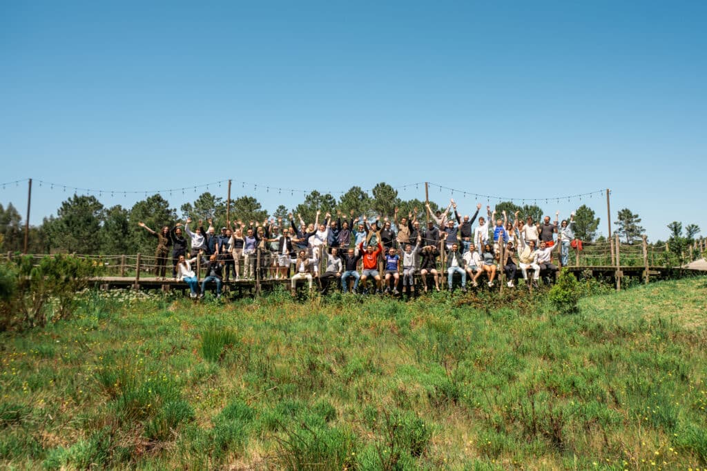 centre of image is a wooden walkway raised above grass on which 50 people are sitting and standing smiling with arms up and bright blue sky