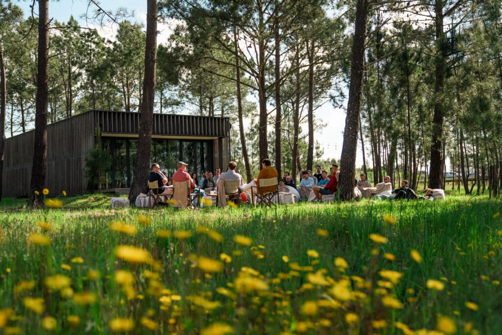 Woodland scene with yellow flowers in foreground, a wooden structure and people sitting on chairs in a circle