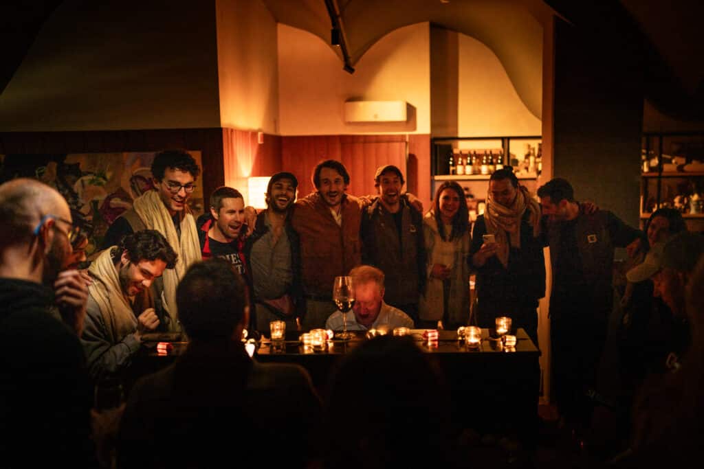 People with arms around each others shoulders smiling and singing around a man playing the piano
